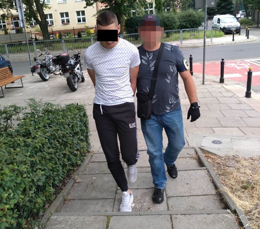 Policja aresztuje podejrzanego, ma założone ręce i jest eskortowany przez funkcjonariusza.