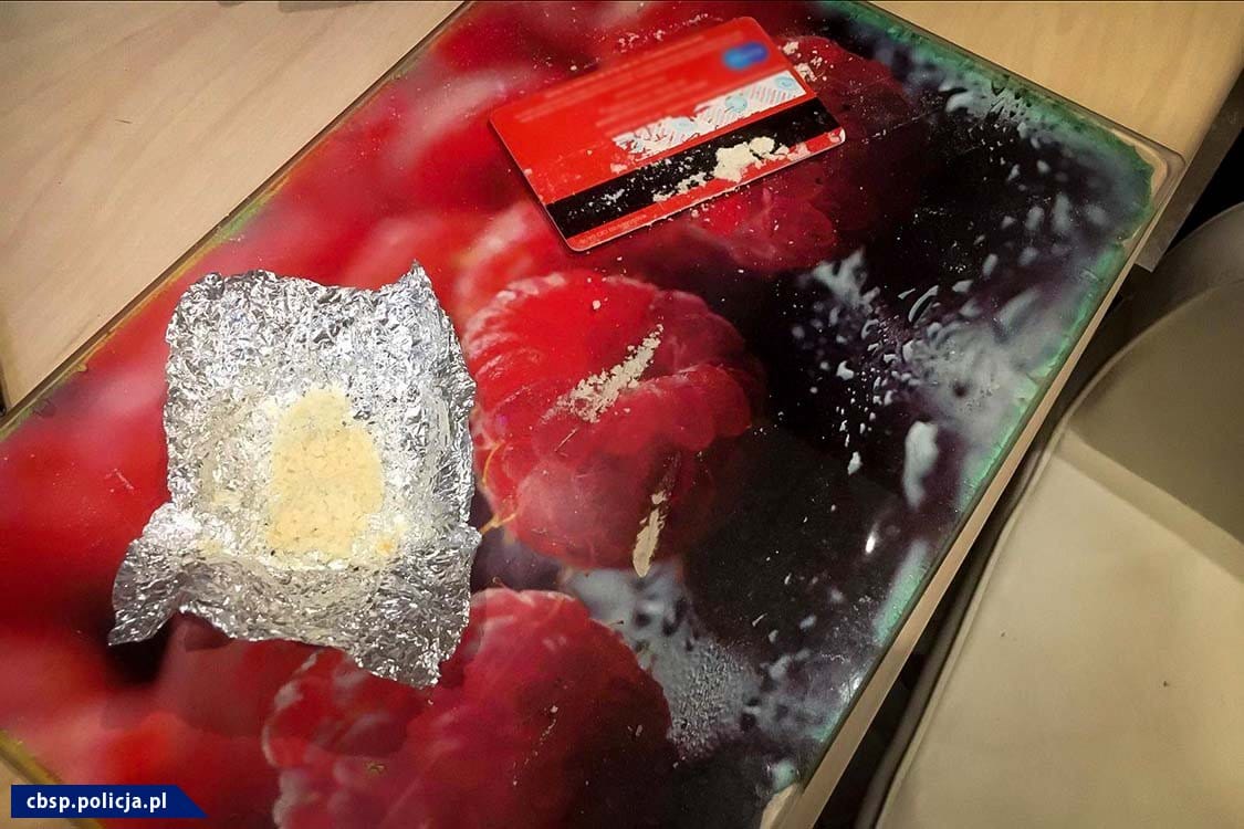 Zbliżenie szklanej powierzchni z kupką białego proszku, foliowym opakowaniem proszku i czerwoną kartą kredytową. Przybory do zażywania narkotyków.