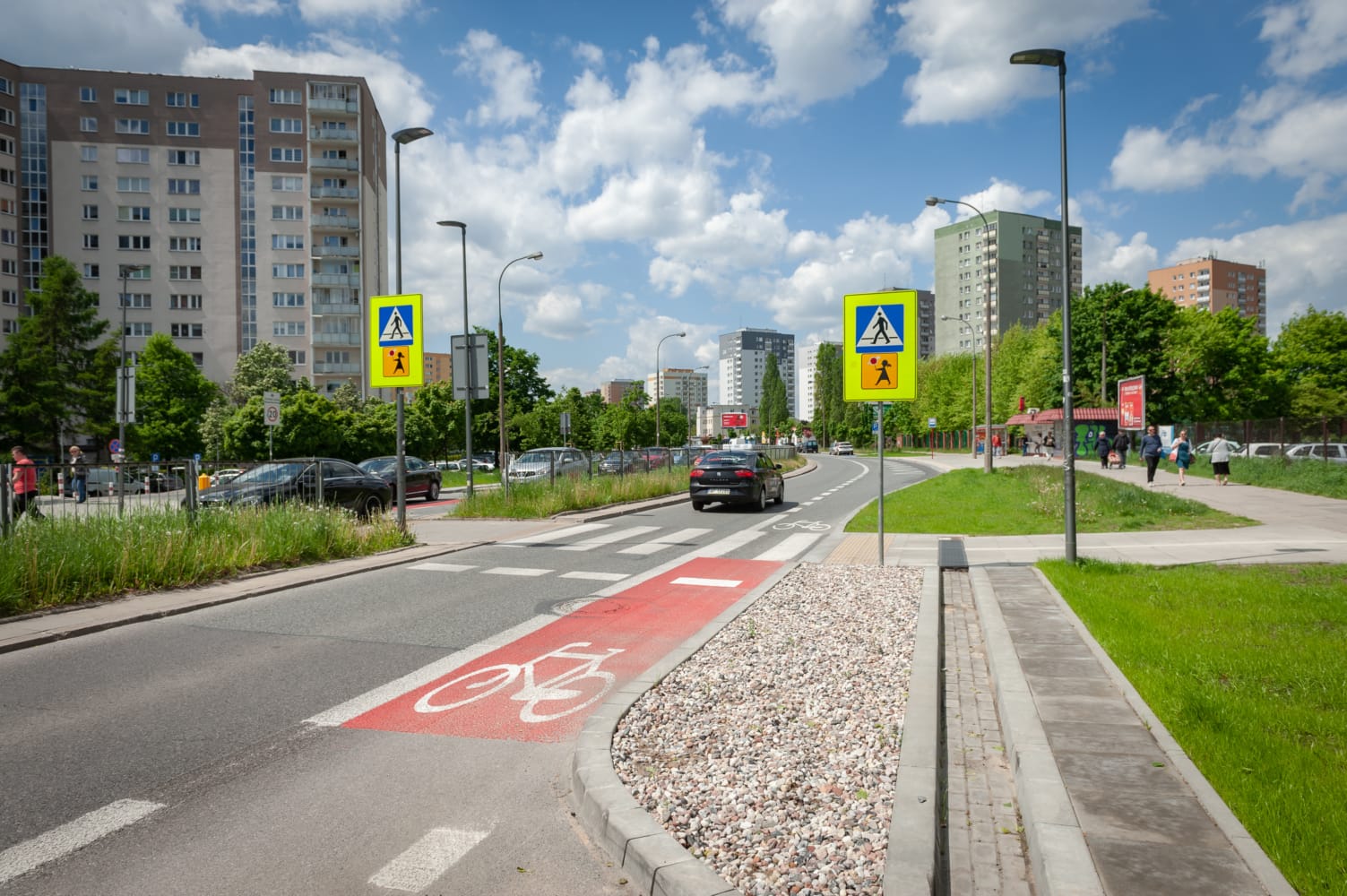 Przejście dla pieszych ze ścieżką rowerową i znakami przejścia dla pieszych na terenie osiedla mieszkaniowego.