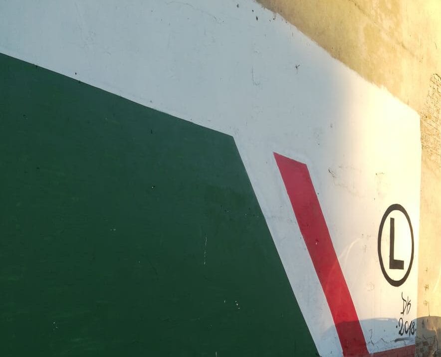 Abstrakcyjna sztuka ścienna z zielonymi, białymi i czerwonymi kształtami geometrycznymi; widoczne logo „L”.