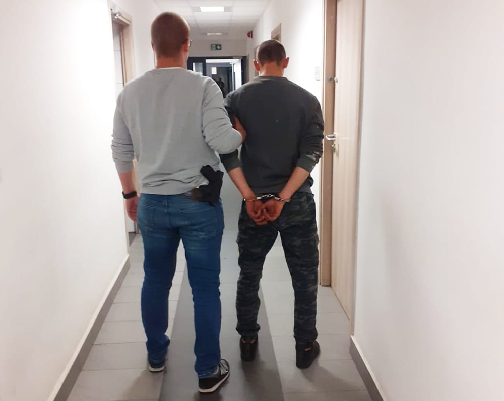 Policjant eskortujący skutego kajdankami podejrzanego korytarzem.