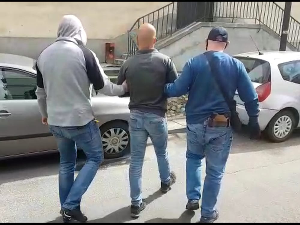 Trzech mężczyzn ubranych w dżinsy i codzienne ubrania, w tym jeden w kajdankach, eskortowanych przez funkcjonariuszy organów ścigania.