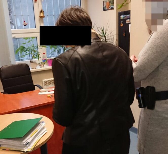 Kobieta w czarnej skórzanej kurtce w biurze rozmawia z osobą ubraną na szaro.