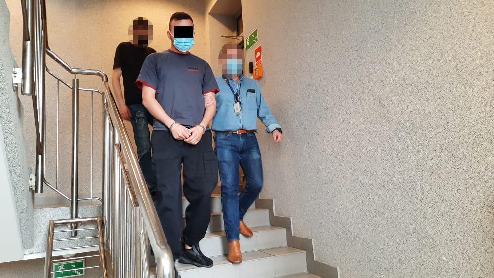 Dwóch mężczyzn w kajdankach eskortowanych po schodach przez policjanta.