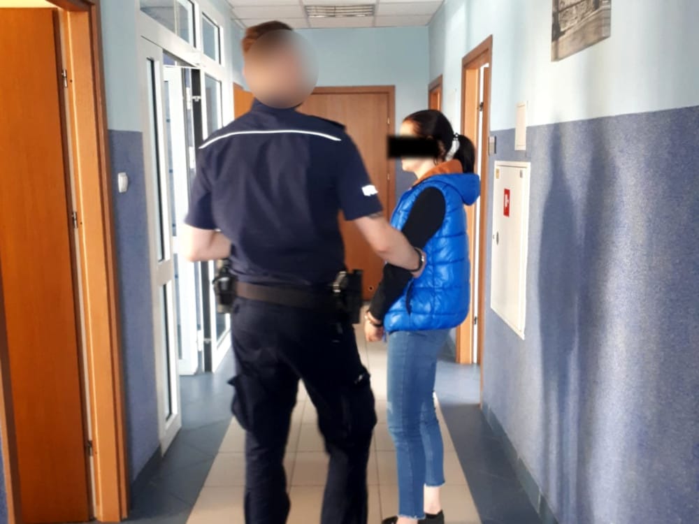 Policjant eskortujący skutą kajdankami kobietę korytarzem.