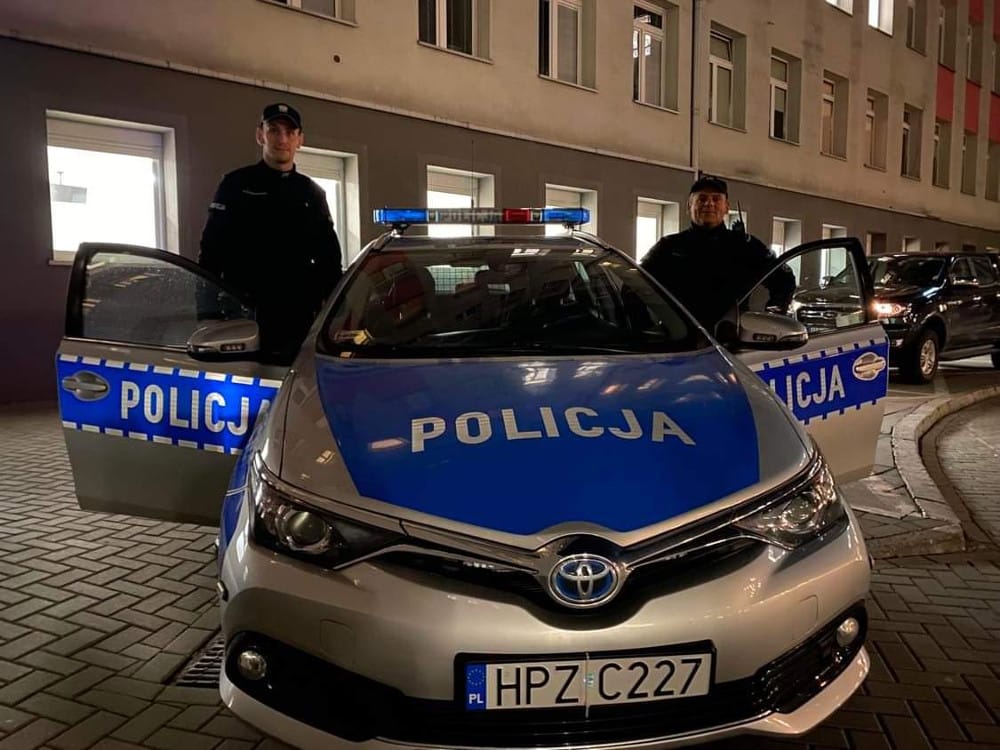 Dwóch polskich policjantów stoi dumnie przy swoim radiowozie z napisem „POLICJA”.
