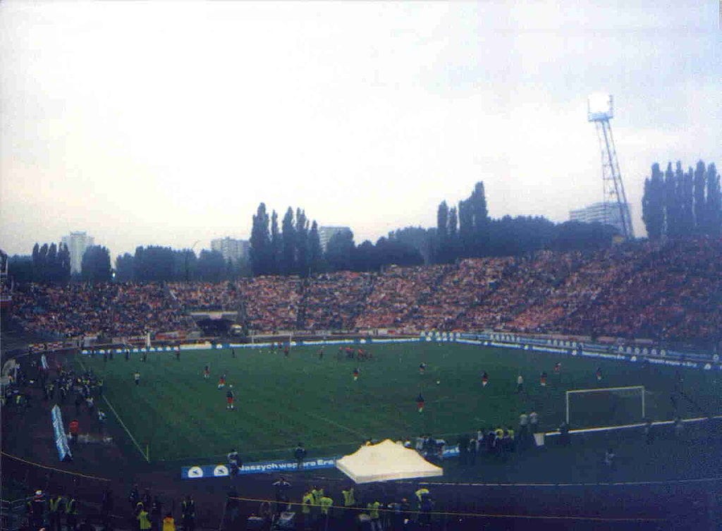 Pełny stadion widzów oglądających mecz piłki nożnej. Boisko jest zielone, a trybuny pełne ludzi.