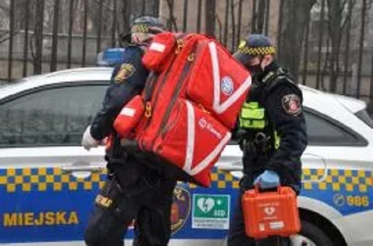 Dwóch ratowników medycznych z torbą medyczną i defibrylatorem obok radiowozu.