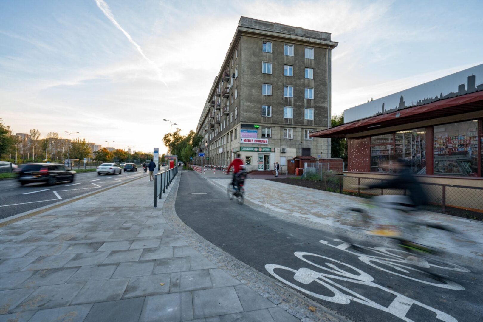 Rowerzyści jadą nową ścieżką rowerową obok ulicy w Warszawie, Polska. Widoczny sklep „Mebłe Biurowe”.