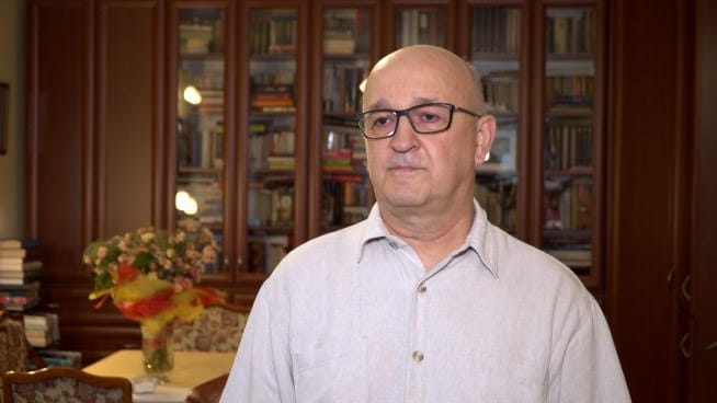 Portret mężczyzny w okularach przed regałem z książkami.