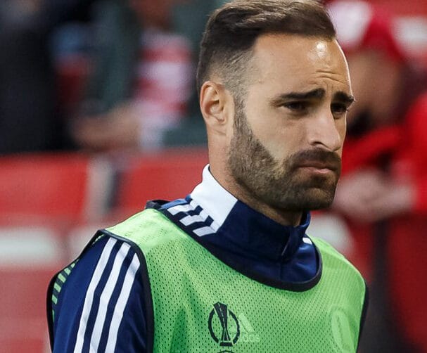 Smutno wyglądający piłkarz ubrany w zielony strój kąpielowy i ciemnoniebieską kurtkę Adidas.