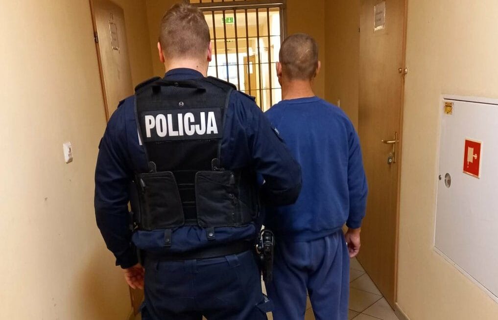 Policjant eskortujący więźnia korytarzem. Na kamizelce policjanta widnieje napis „POLICJA.