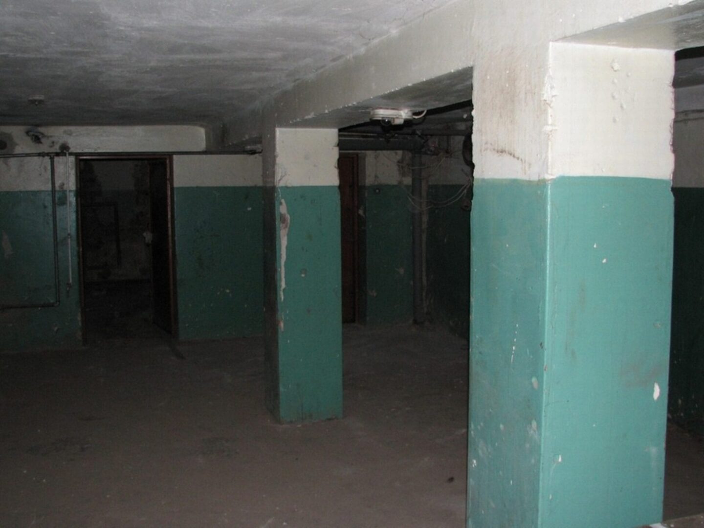 Ciemna, opuszczona piwnica z łuszczącą się turkusową farbą na kolumnach podporowych.