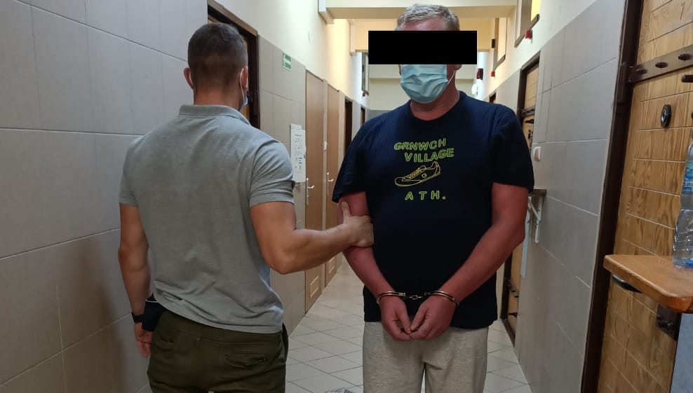 Policjant eskortujący zakutego w kajdanki podejrzanego korytarzem. Podejrzany ma na sobie koszulkę z napisem „Greenwich Village ATH”.