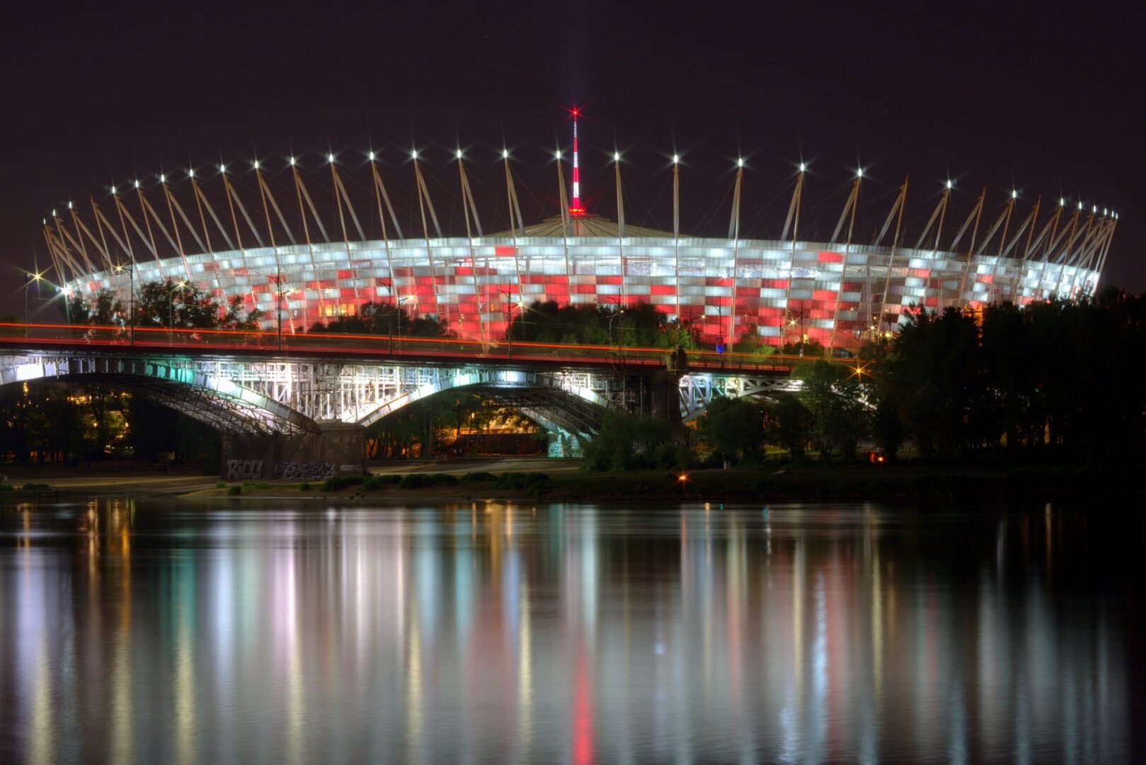 Nocny widok na oświetlony Stadion Narodowy w Warszawie, odbijający się w Wiśle.