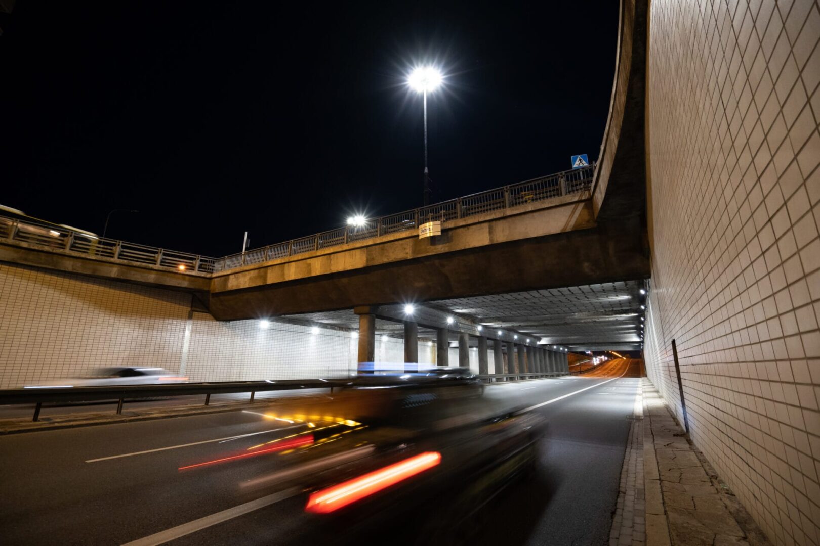 Nocny widok samochodów przejeżdżających przez przejazd podziemny pod autostradą, oświetlony latarniami ulicznymi.
