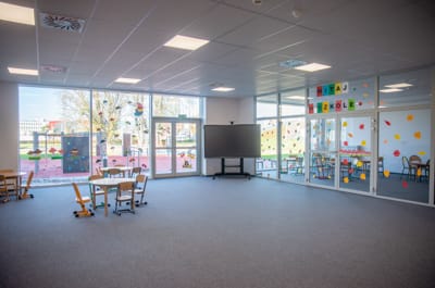 Przestronna sala lekcyjna ze stołami, krzesłami i interaktywną tablicą w rozmiarze dziecięcym. Duże okna zapewniają naturalne światło.