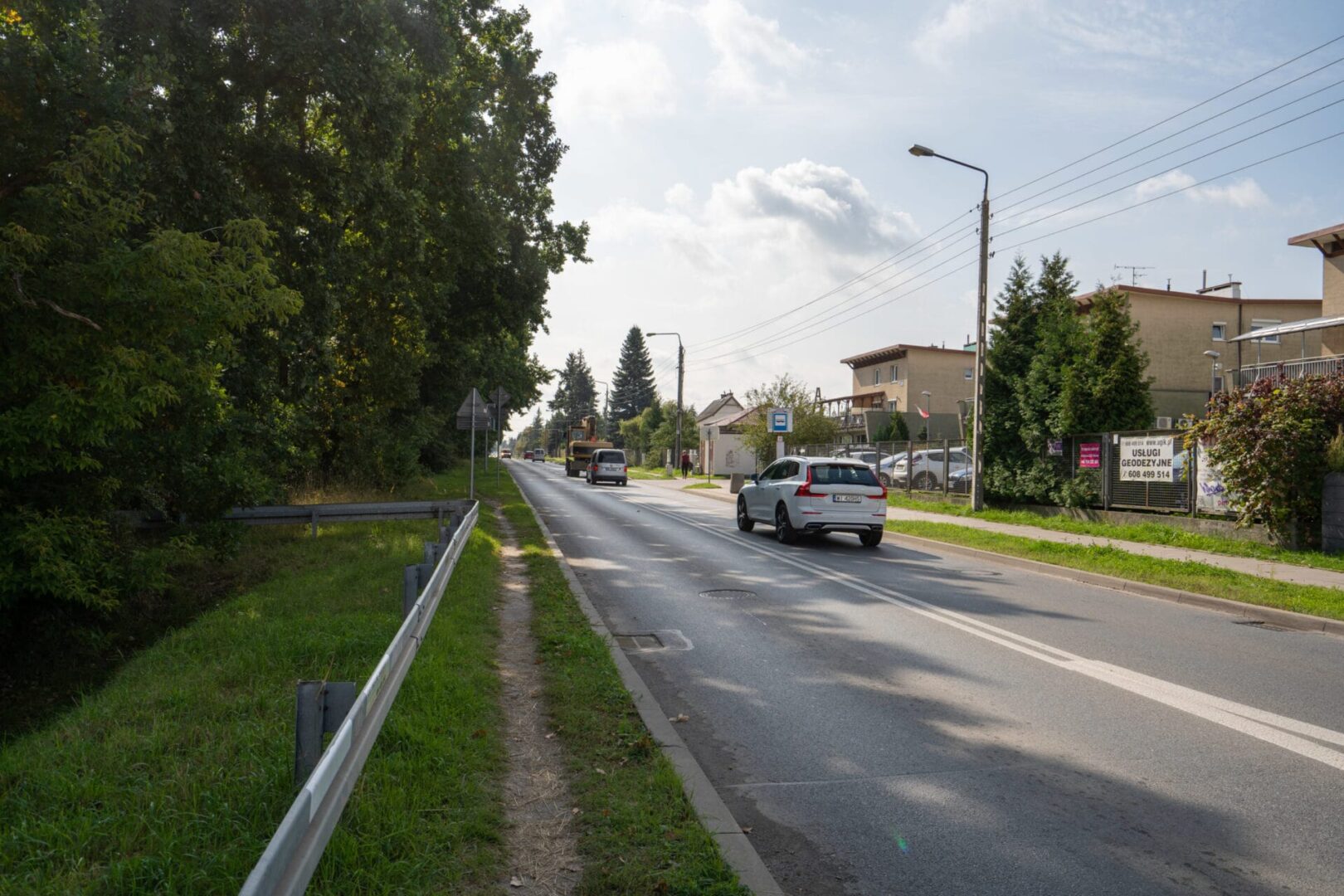 Samochody jadące drogą osiedlową otoczoną drzewami i domami.