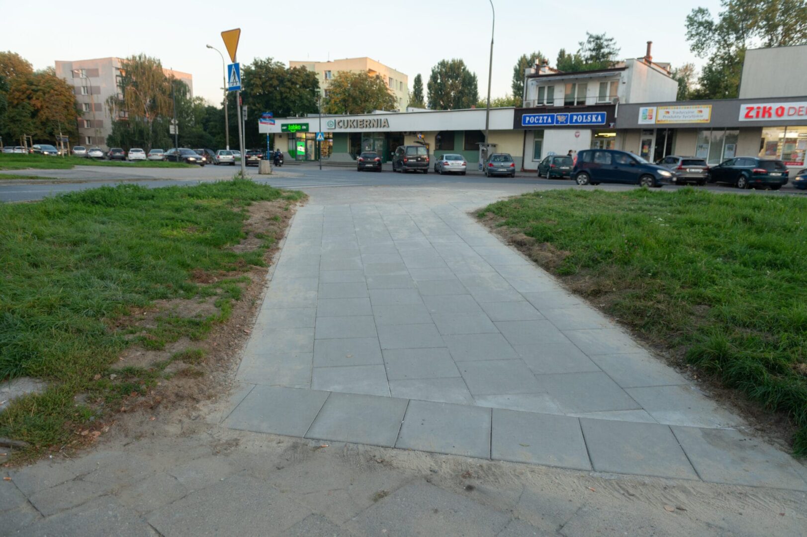 Nowo wyasfaltowane przejście dla pieszych w pobliżu sklepów, w tym cukierni i poczty polskiej.