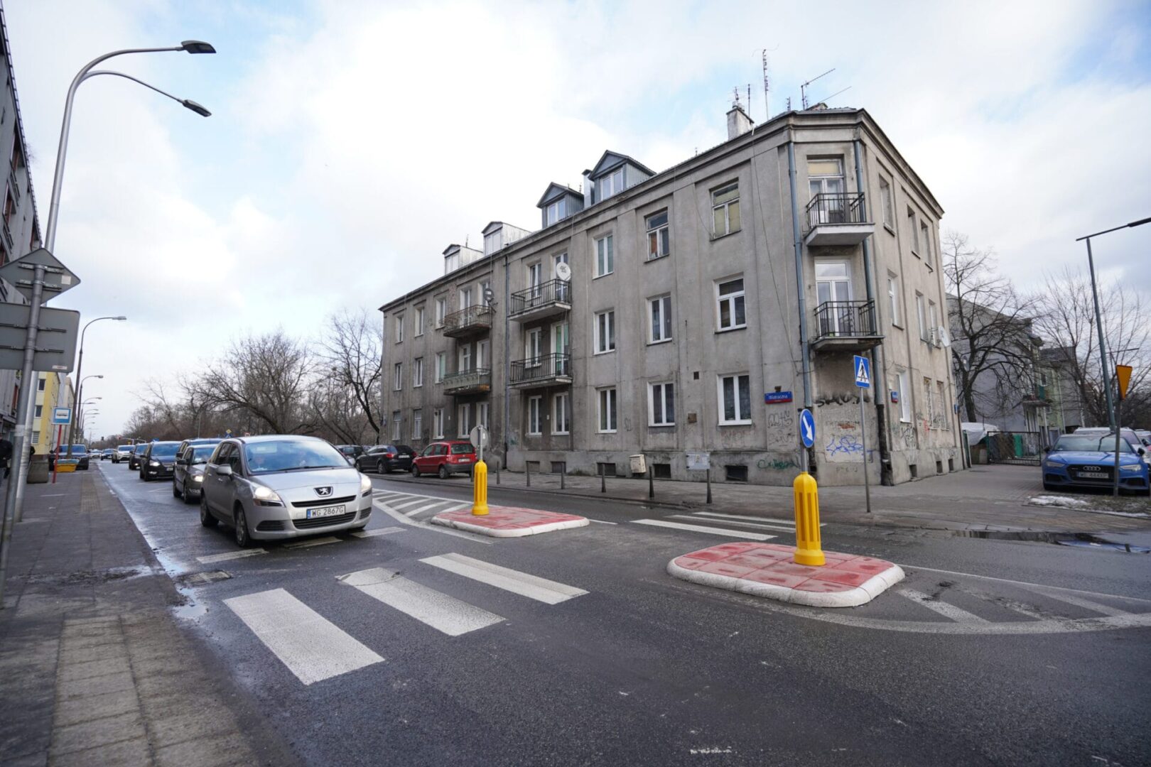 Samochody przejeżdżające obok przejścia dla pieszych w pobliżu szarego budynku mieszkalnego w Warszawie.