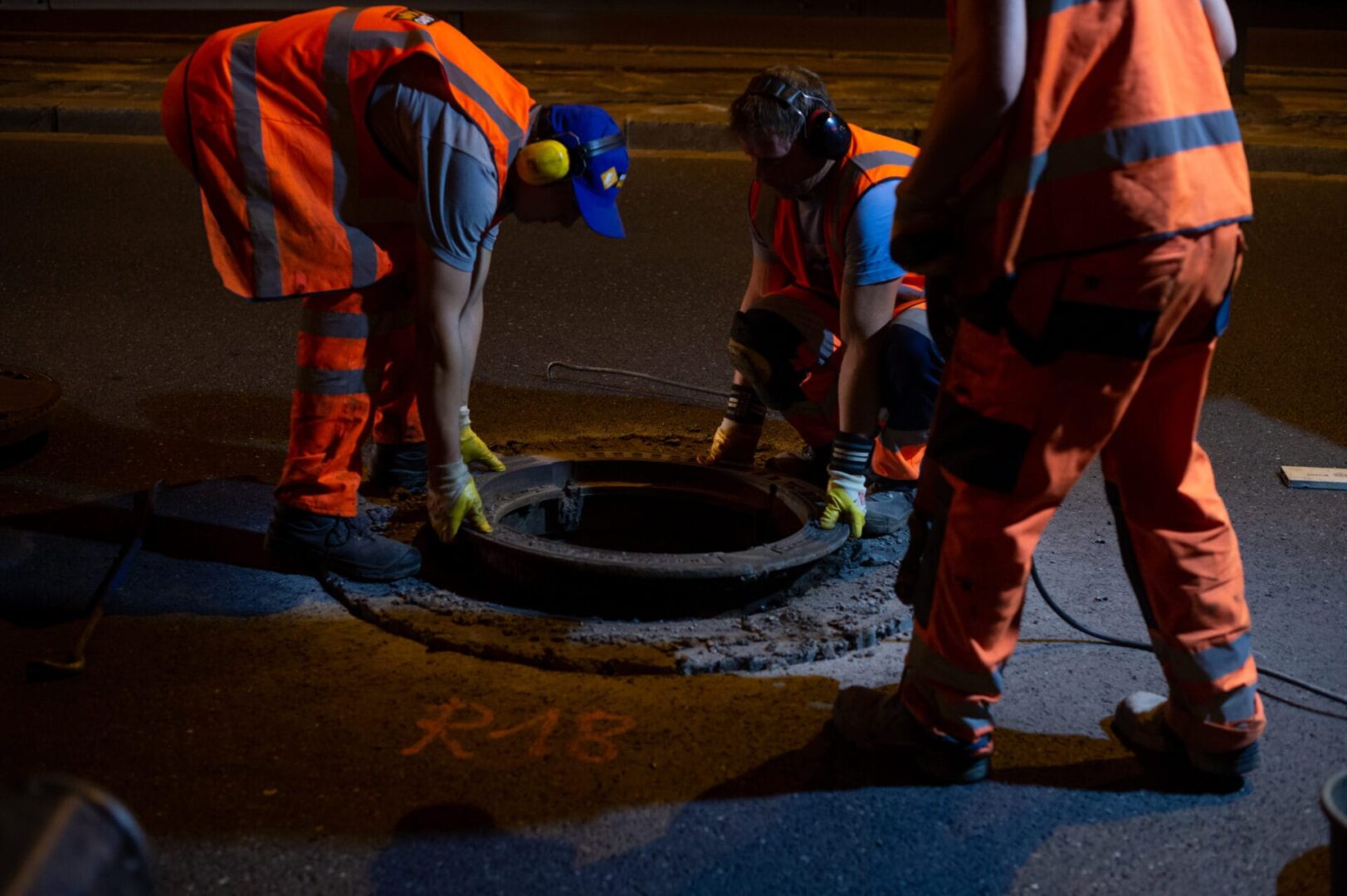 Trzech pracowników w kamizelkach odblaskowych wymienia pokrywę włazu studzienki w nocy.