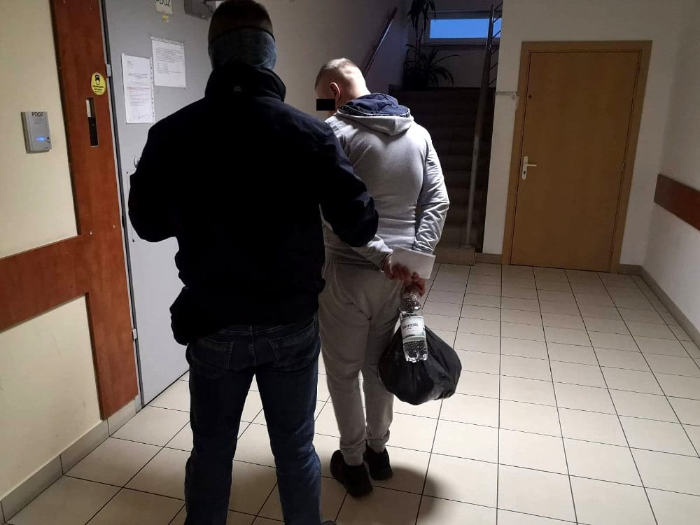 Policjanci eskortujący podejrzanego, mającego skute ręce i torbę.