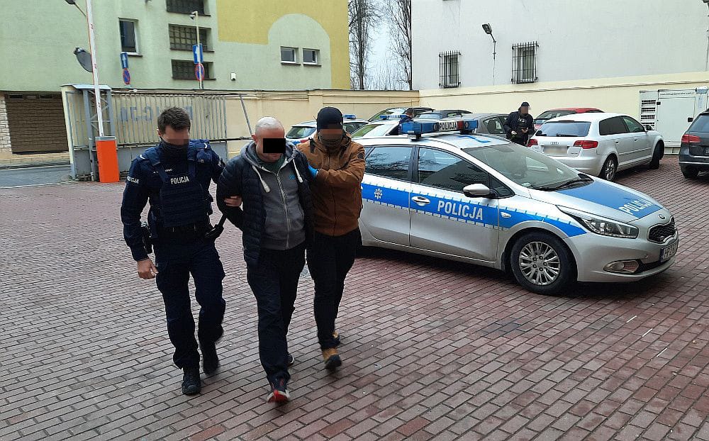 Policjanci aresztujący podejrzanego. Na mundurach policjantów i radiowozie widać napis „Policja”.