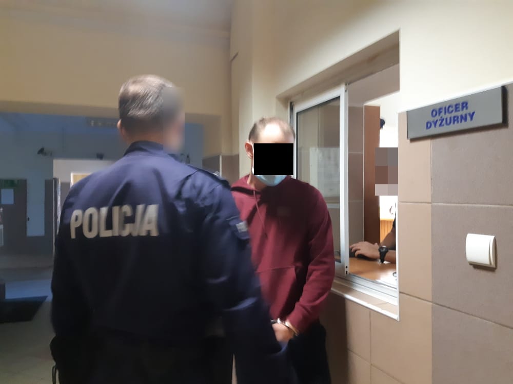 Policjant eskortujący podejrzanego w kajdankach na posterunku policji. Na mundurze policjanta widoczny jest napis „POLICJA”.