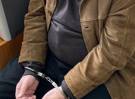 Zbliżenie dłoni mężczyzny w kajdankach, ubranego w brązową zamszową kurtkę.