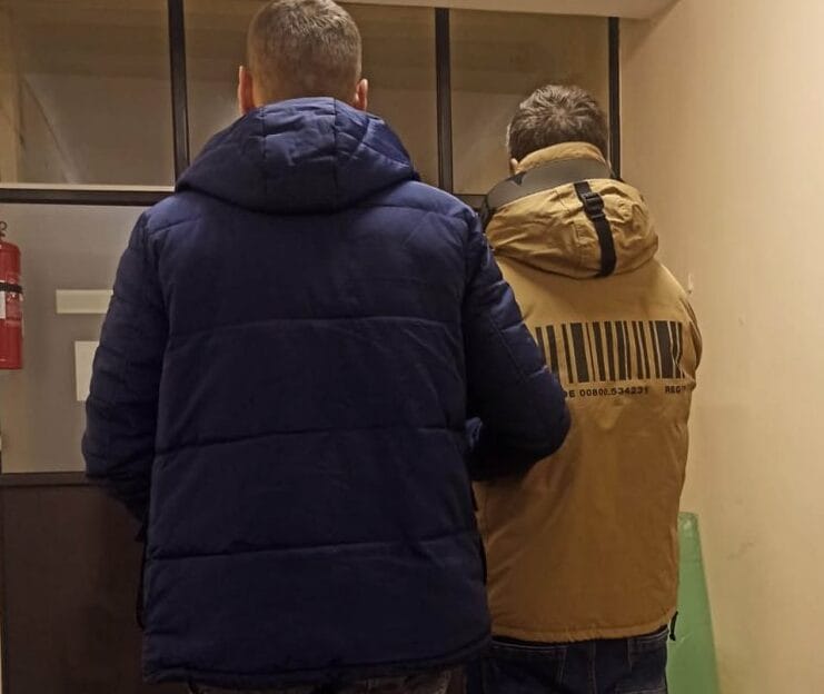 Dwóch mężczyzn w zimowych kurtkach stoi na korytarzu. Jedna kurtka jest granatowa, druga jasnobrązowa z nadrukowanym kodem kreskowym na plecach.