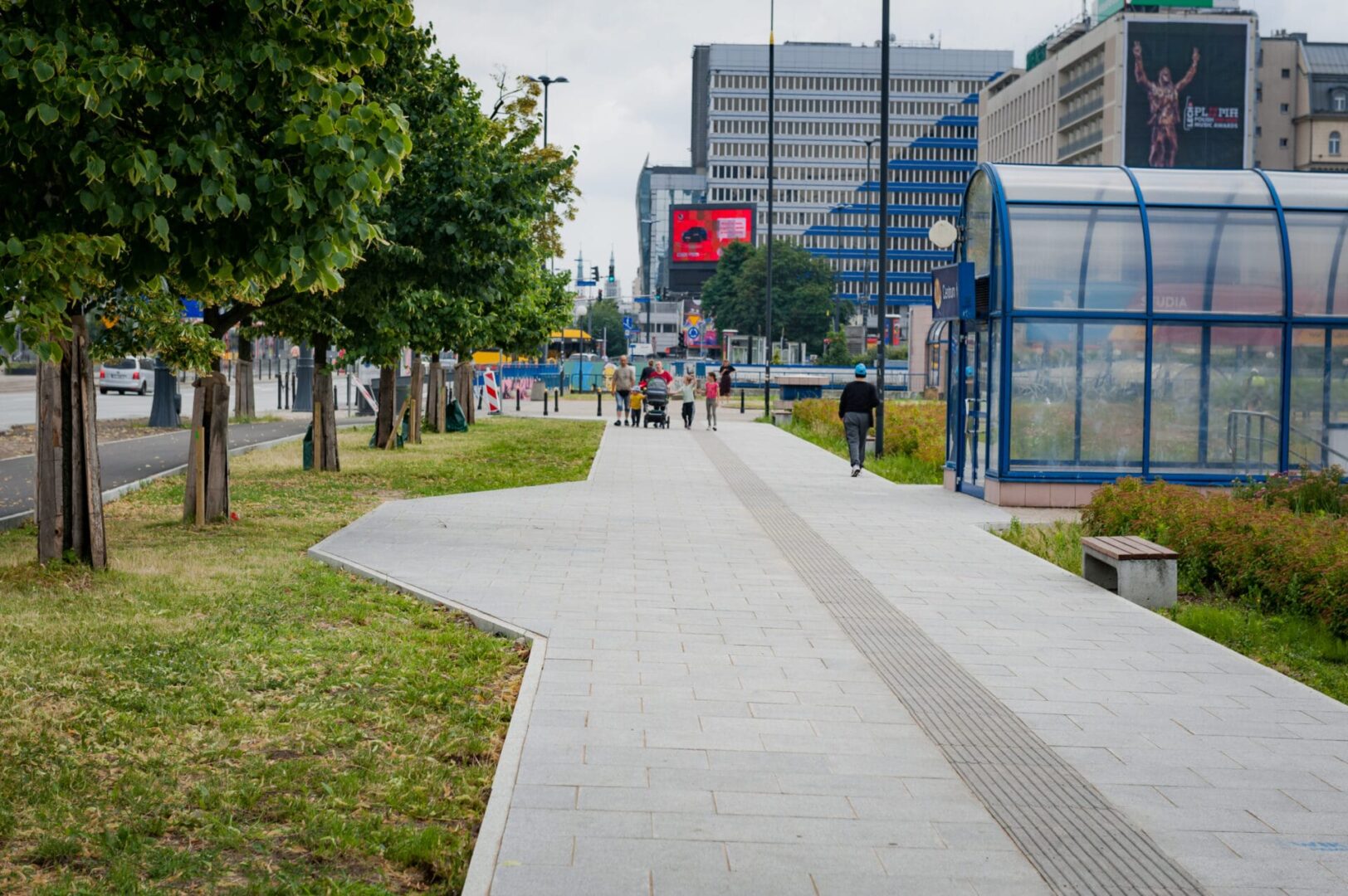 Kładka dla pieszych z ludźmi, drzewami i nowoczesnymi budynkami w tle.