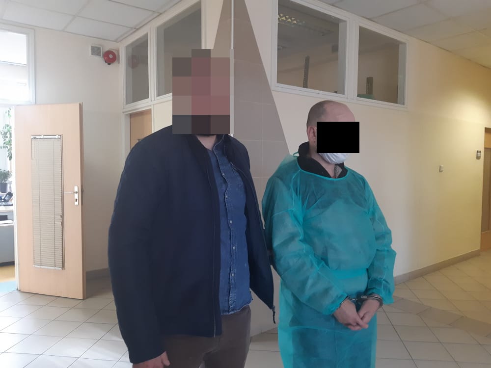 Dwóch mężczyzn w areszcie, jeden ubrany w niebieską kurtkę, drugi w turkusowy fartuch i kajdanki.