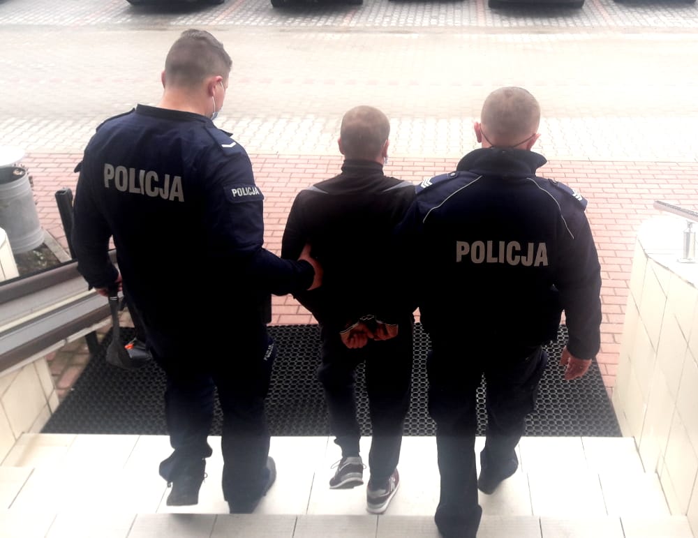 Dwóch polskich policjantów eskortuje skutego kajdankami podejrzanego po schodach. Na ich mundurach widać napis POLICJA.